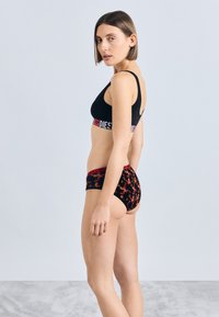 Černá braletka se širokým červeno-bíl�ým páskem s logem, kombinovaná s černým spodním prádlem s červeným květinovým vzorem a sladěným pasem.