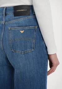 Mėlynos džinsinės kelnės su šiek tiek nusidėvėjusiu tekstūru, turinčios juodą odinę etiketę ant juosmens ir auksinę erelio logotipą ant užpakalinio kišenės.