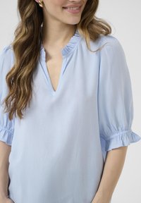 Blusa azul claro con cuello en V y detalles de volantes. Las mangas cortas presentan acentos de volantes gather. Tela suave con un ligero brillo.