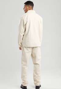 Beige denim jacket with buttoned cuffs, relaxed fit, and matching beige pants, set against a simple gray background. Black shoes visible.