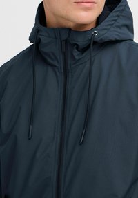 Chaqueta de color gris oscuro con cremallera frontal, cuello alto y ajuste con cordones, además de una textura suave y mate. No presenta logotipos visibles.