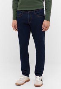 Granatowe dżinsy z ciemnoniebieskiego denimu z kontrastującym pomarańczowym przeszyciem, pięciokieszeniowym fasonem i prostą nogawką, zestawione z kremowymi trampkami.