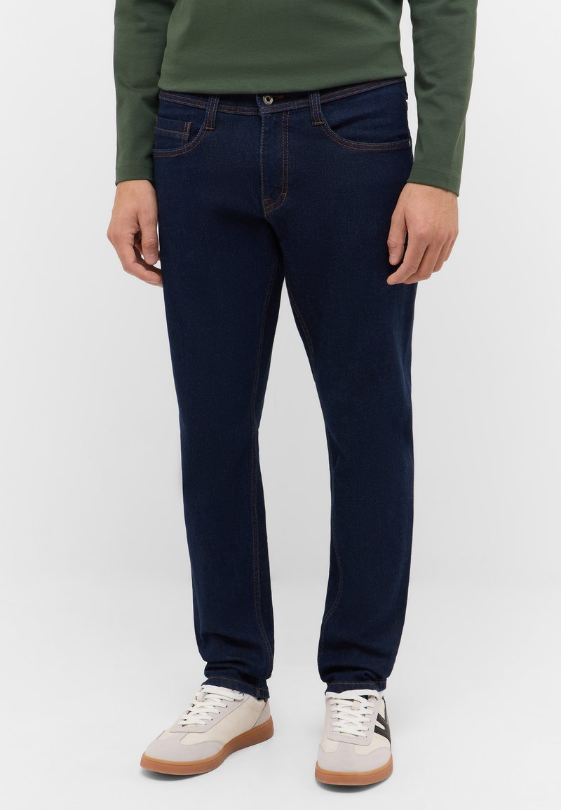 Granatowe dżinsy z ciemnoniebieskiego denimu z kontrastującym pomarańczowym przeszyciem, pięciokieszeniowym fasonem i prostą nogawką, zestawione z kremowymi trampkami.