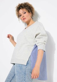 Sudadera gris con escote en V y paneles laterales a rayas azules. Con mangas largas ligeramente abullonadas y un corte relajado. Parte inferior de mezclilla casual.