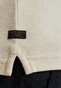 Tissu texturé beige avec un motif tissé, doté d'une étiquette noire avec un texte doré, située à l'arrière près de l'ourlet avec un détail découpé.