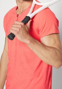 Camiseta atlética de coral brillante con mangas cortas, tejido texturizado y cuello con cremallera, sostenida con una mano que agarra una raqueta de tenis.