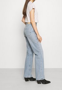 Nainen seisoo sivuttain valkoista taustaa vasten, päällään vaaleansiniset korkeavyötäröiset suoralahkeiset farkut, valkoinen lyhythihainen paita ja mustat korolliset nilkkurit.