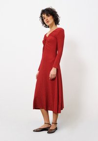 Rotes langärmliges Kleid mit tiefem V-Ausschnitt und gerafftem Vorderteil, fließender Rock, aus strukturiertem Stoff. Kombiniert mit schwarzen Netz-Ballettschuhen.