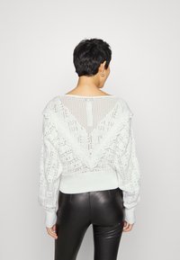 Pull blanc en maille avec motifs texturés et détail dos ouvert. Présente un large décolleté et des poignets côtelés, associé à un pantalon noir.