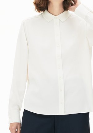 Femme portant une chemise blanche à manches longues avec col et boutons, et un pantalon foncé, ajustant ses cheveux d'une main.