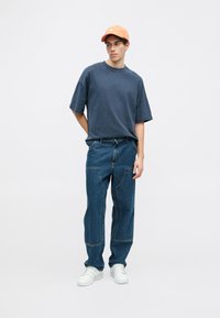 Mladý muž v oranžové čepici, volném tmavě modrém tričku, pohodlných džínách s náprsními kapsami a bílých teniskách stojící na prostém bílém pozadí.