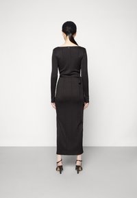 Robe noire à manches longues côtelée avec une silhouette cintrée, un décolleté carré et une ceinture à nouer à la taille. La jupe arrive au milieu du mollet.