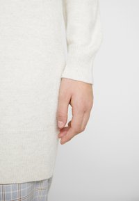 Höger hand avslappnad vid sidan, klädd i en krämfärgad stickad tröja och ljusgrå rutig byxa mot en enfärgad bakgrund.