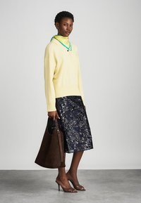 Gelbes Langarmoberteil, marineblumiger Rock, braune Wildlederhandtasche und braune High-Heels. Heller Hintergrund, Fokus auf Kleidung.