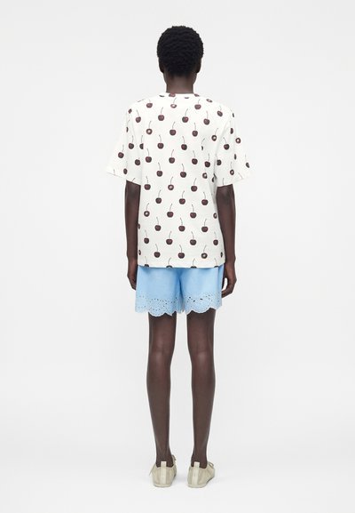 Άτομο στέκεται πλάτη φορώντας λευκό πουκάμισο με σχέδια κερασιών, ανοιχτό μπλε σορτς με κυματιστό τελείωμα και μπεζ παπούτσια χωρίς κορδόνια.