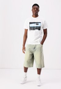 Camiseta blanca de algodón con un estampado gráfico del interior de un coche; combinada con pantalones cortos de mezclilla verde claro y zapatillas blancas con calcetines cortos.