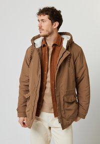 Parka isolata marrone con cappuccio foderato in pelliccia bianca, chiusura con zip e tasche frontali. Indossata sopra una camicia di velluto a coste color ruggine e strati beige.
