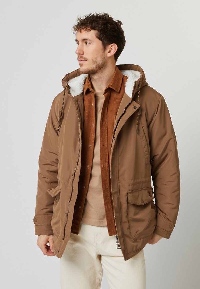 Parka isolata marrone con cappuccio foderato in pelliccia bianca, chiusura con zip e tasche frontali. Indossata sopra una camicia di velluto a coste color ruggine e strati beige.