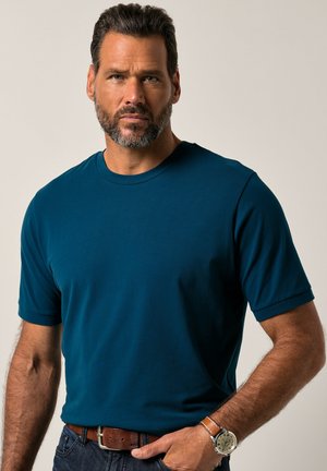 Camiseta de manga corta con cuello redondo en teal profundo, fabricada en algodón suave. Combinada con un cinturón y un reloj marrones, presenta un corte relajado y un color sólido.