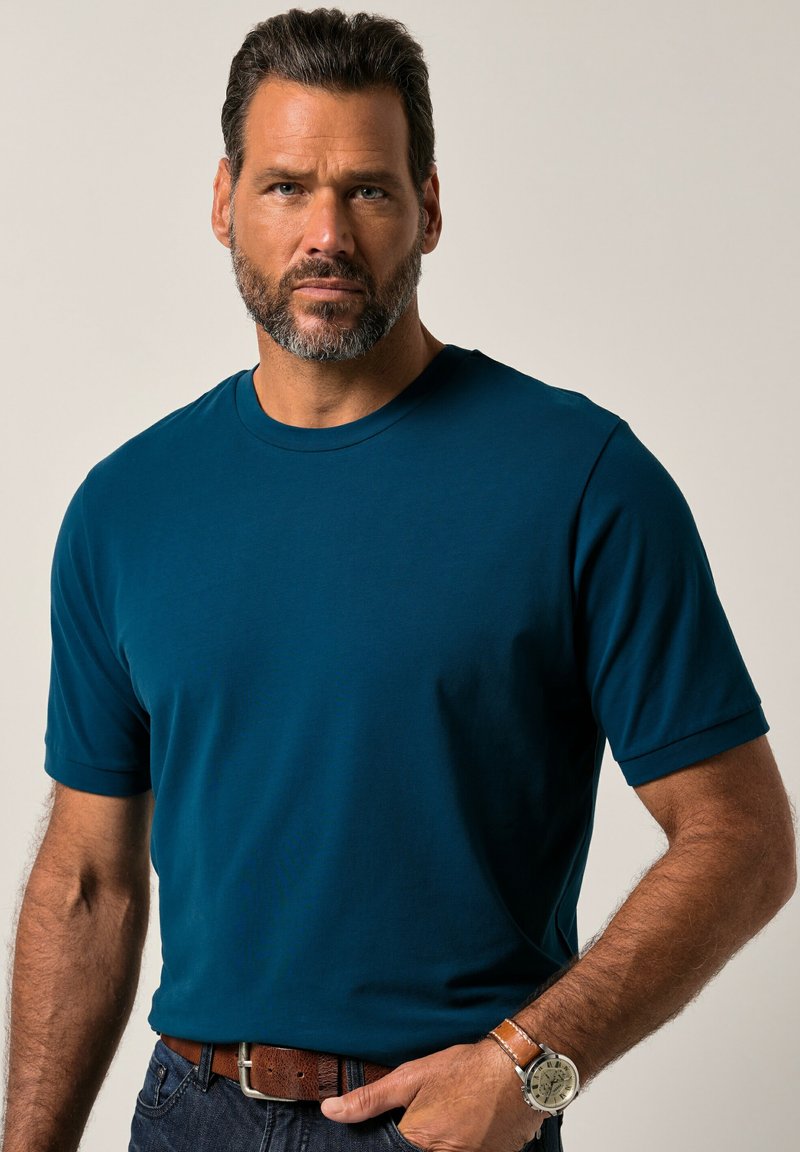 T-shirt a maniche corte con scollo rotondo in un profondo teal, realizzato in cotone liscio. Indossato con una cintura e un orologio marroni, presenta una vestibilità rilassata e un colore uniforme.
