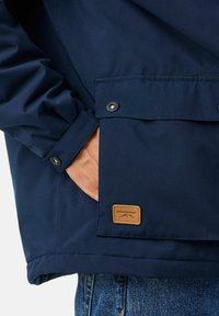 Marine waterproof jack met drukknoopsluiting, een voorzak en een bruine leren logo patch. Materiaal lijkt glad en duurzaam.