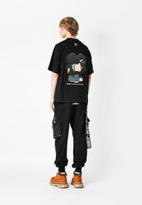 Camiseta gráfica negra con un colorido estampado en la parte posterior, combinada con pantalones cargo negros con bolsillos laterales y botas marrones. Estilo casual.