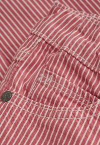Rot-weißer gestreifter Stoff, der eine Nahaufnahme von genähten Kanten und einem metallischen Knopfdetail in der Nähe einer Tasche zeigt. Die Textur wirkt weich.