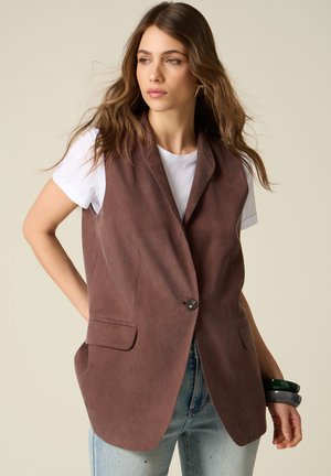 Donna che indossa un blazer marrone senza maniche sopra una t-shirt bianca e jeans azzurri, tenendo due braccialetti al polso.