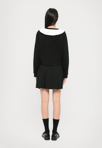 Maglione nero a maglia con colletto a coste bianco, abbinato a una gonna nera plissettata. Il modello indossa calze nere fino al ginocchio e scarpe con fibbie.