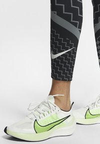 Białe buty sportowe z zielonymi akcentami i czarnymi detalami, cechujące się lekką konstrukcją i oddychającymi panelami z siateczki. Czarn leginsy w wzory.