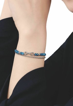 Handgelenk mit zwei Armbändern: eines mit blauen Perlen und silbernem Unendlichkeitssymbol sowie eine dünne silberne Kette, vor einem schwarzen Ärmel.