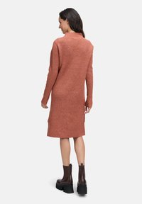 Vrouw met haar rug naar de camera, gekleed in een geribbelde, langmouwige, roestkleurige midi-pulloverjurk en donkere bruine chunky platform enkellaarzen.