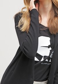 Donna bionda che indossa un cardigan nero e una t-shirt nera con una silhouette grafica bianca di una persona che indossa occhiali da sole.