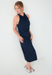 Femme en robe sans manches bleu marine à col halter et talons ouverts bleu clair, debout contre un mur blanc, regardant sur le côté.