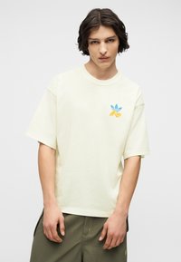 Νεαρός άνδρας με μεσαίου μήκους σκούρα μαλλιά, φορόντας ένα μπεζ μπλουζάκι με μπλε και κίτρινο λογότυπο στο στήθος και παντελόνι ελιάς.