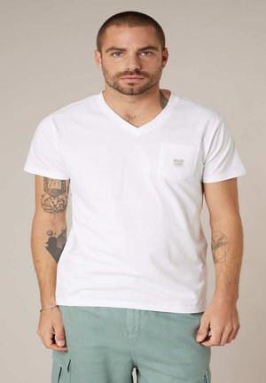 Man met kort haar en baard, gedragen een witte V-hals T-shirt met borstzak en lichtgroene cargobroek, staand tegen een neutrale achtergrond.