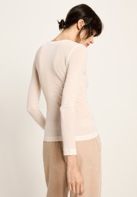 Frau mit dunklem, nach hinten gebundenem Haar, die ein enganliegendes, cremefarbenes Langarmoberteil und beigen strukturierten Hosen trägt, seitlich vor einem schlichten Hintergrund.