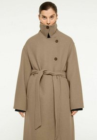 Beige ullblandad kappa med hög krage, dubbelknäppta knappar och bälte. Har vida ärmar och stora sidofickor.