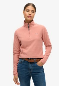 Pullover in pile rosa con colletto a mezza zip, dotato di un piccolo logo sul petto. Abbinato a jeans blu e una cintura marrone. Texture liscia.