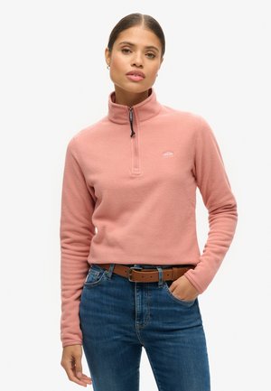 Pullover in pile rosa con colletto a mezza zip, dotato di un piccolo logo sul petto. Abbinato a jeans blu e una cintura marrone. Texture liscia.