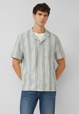 Jonge man die een kortemouwen overhemd met verticale grijze en witte strepen en een blauwe spijkerbroek draagt, die tegen een effen grijze achtergrond staat.