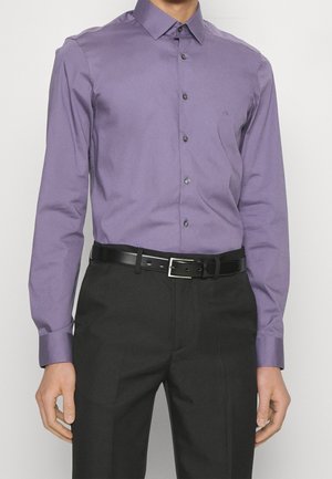 Hombre con una camisa morada ajustada metida dentro de pantalones negros con un cinturón de cuero negro, de pie frente a un fondo liso.