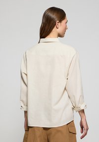 Jasno beżowa, oversize koszula z karczkiem z tyłu, rękawami na zapięcie oraz kołnierzykiem, wykonana z teksturowanej tkaniny.