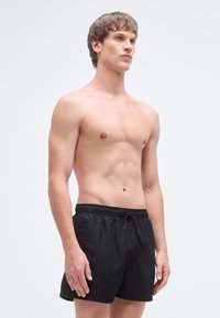 Juodos spalvos plaukimo šortai su elastiniu juosmeniu ir raištelio detalėmis, pasižymintys lygiu paviršiumi ir plokščiomis siūlėmis. Modelis stovi be marškinėlių.