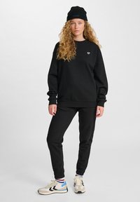 Giovane donna con lunghi capelli ricci, che indossa un berretto nero, una felpa nera, pantaloni neri e sneakers bianche con dettagli blu e neri.