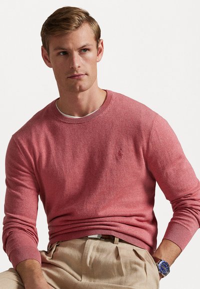 Rožinė megzta palaidinė su apvalia kaklo iškirpte ir raukšlėtomis rankovėmis, derinama su rusvom kelnėmis. Suknelėje yra mažas logotipas ant krūtinės. Lygus tekstūros paviršius.