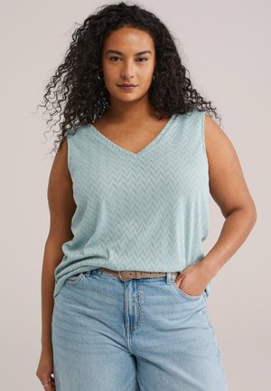 Kvinde med krøllet hår iført en lyseblå mønstret ærmeløs top, stoppet ned i lyseblå jeans med brunt bælte, poserer med den ene hånd i lommen.