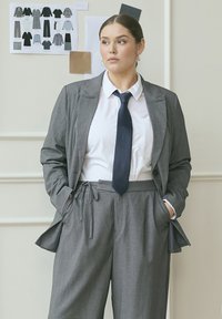 Grå stribede jakkesæt med en tætsiddende blazer og afslappede bukser, parret med en skarp hvid skjorte og navy slips. Glat stof og skræddersyet design.