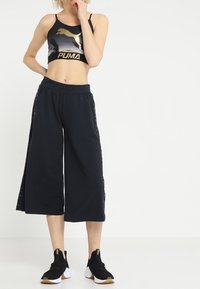 Black sports bra with a metallic gold logo, paired with loose, black cropped pants. Lightweight fabric and black, slip-on athletic shoes with gold accents.