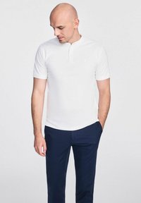 Biała koszulka polo z krótkim rękawem i kołnierzykiem typu henley, w zestawieniu z granatowymi spodniami. Gładka faktura, minimalistyczny design i subtelny detal logo.